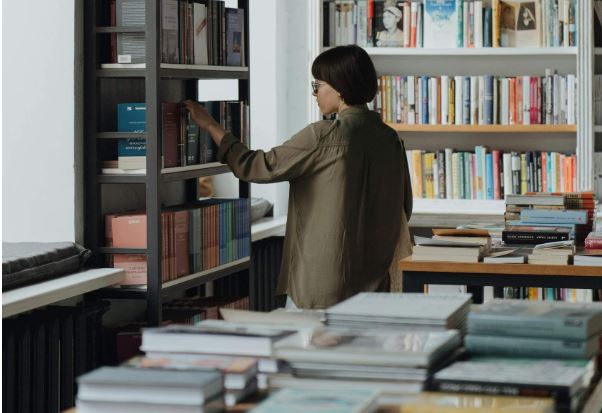 Junge Frau in einer Holzbibliothek und einem Tisch mit vielen aufgeklappten Büchern als Symbol für eine Erleichterung, wenn man alles in einer zentralen Suche findet.