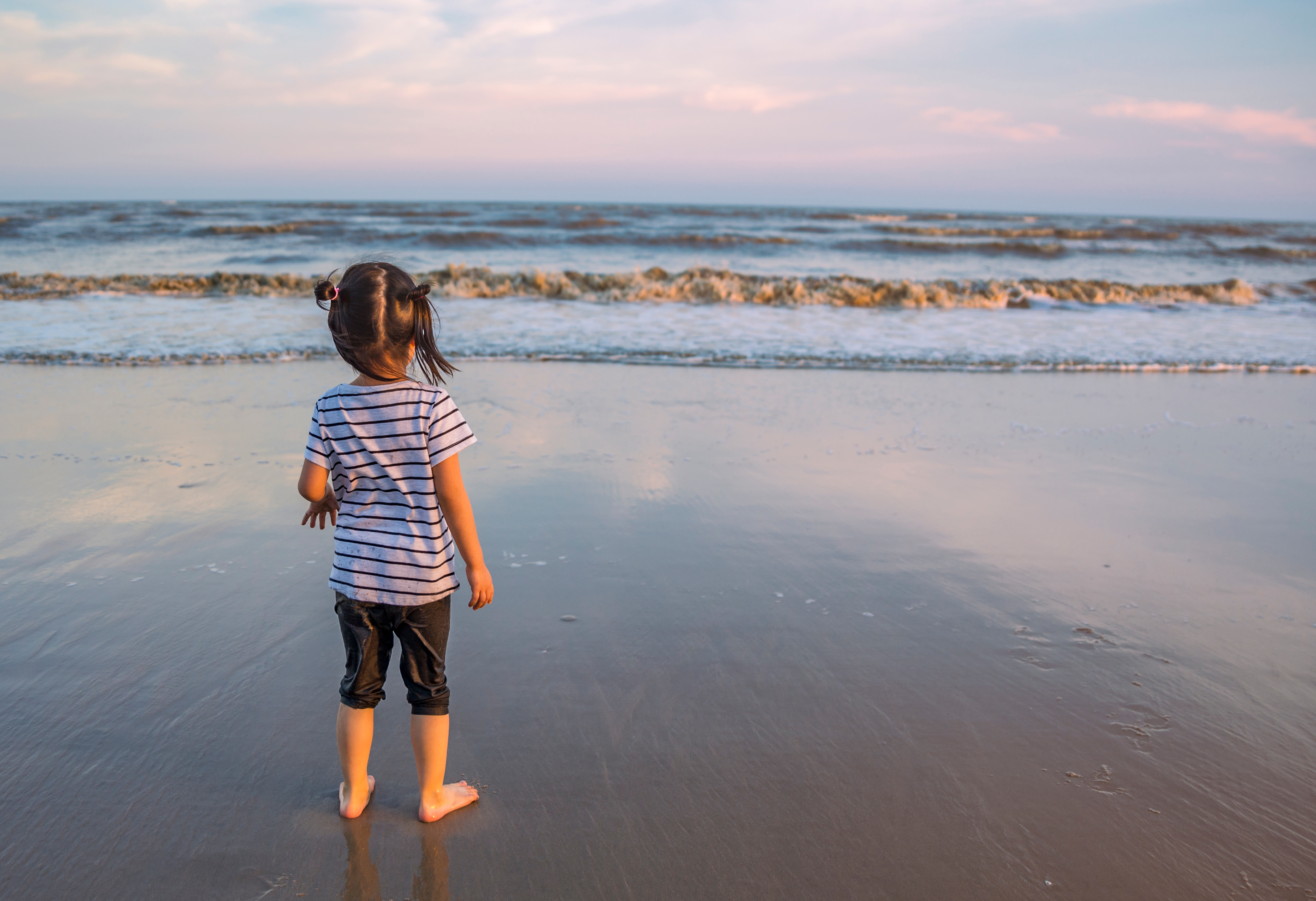 Ein Mädchen mit gestreiftem T-Shirt und dunkler Hose steht barfuß am Strand und schaut auf das Meer. Die Wellen brechen sanft im Hintergrund.