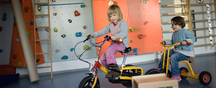 Zwei Kinder fahren auf gelben Dreirädern in einem Raum mit Kletterwand.