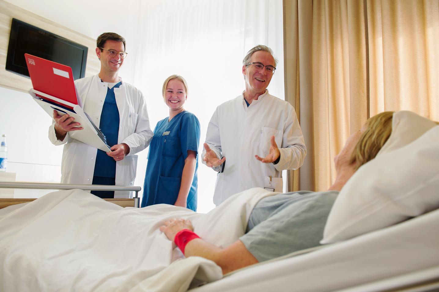 Drei Ärzte und Ärztinnen stehend lachend am Bett einer Kranken. Sie halten dabei Unterlagen in  der Hand