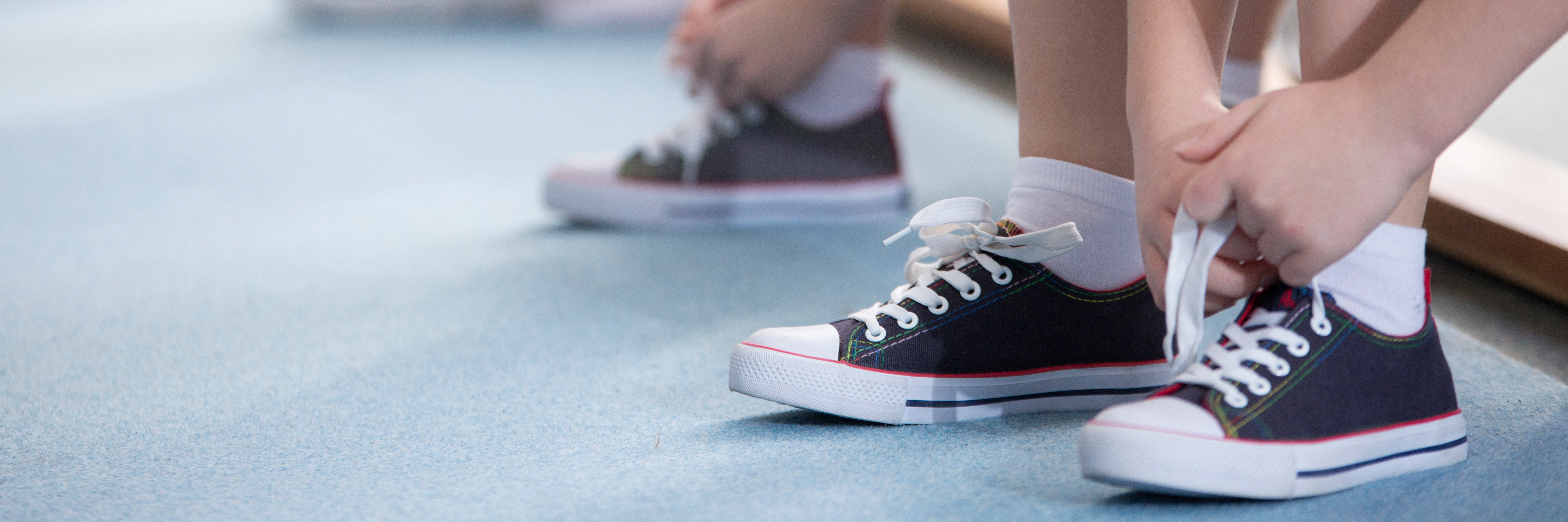 Zwei Hände binden die Schnürsenkel von grauen Sneakers mit weißen Socken auf einem blauen Boden.