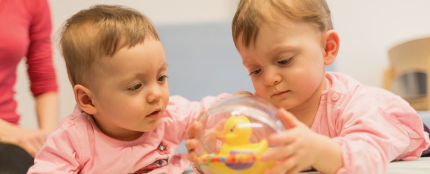 Zwei Kleinkinder spielen mit einer transparenten Kugel, in der eine gelbe Gummiente ist.