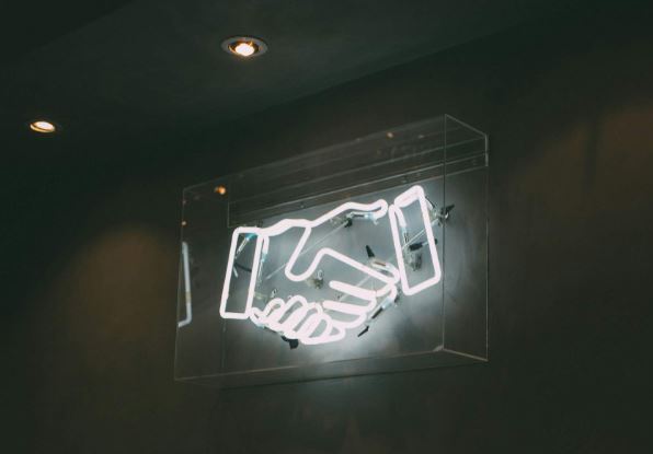 Ein dunkler raum mit weißen stilisierten Neonröhren-Händen, die sich schütteln als Symbol für eine verbesserte Zusammenarbeit durch eine Enterprise Suche.